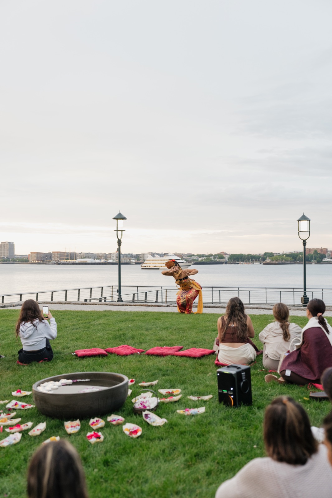 Women seated on the lawn watching a Balinese dancer beside Boston Harbor